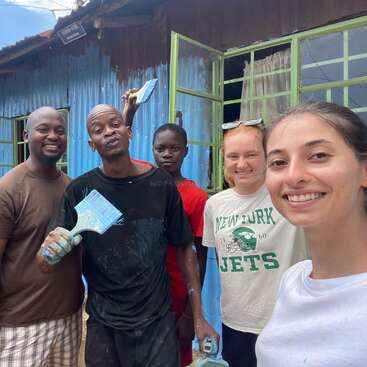 A imagem mostra um grupo de pessoas pintando uma casa, com uma mulher e quatro homens juntos, segurando pincéis e sorrindo para a câmera.