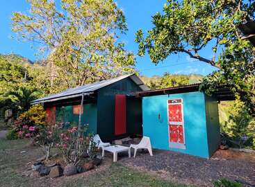 A small, colorful house with turquoise and red accents sits among lush greenery. Two white chairs and a table are placed outside. Bright, sunny day.