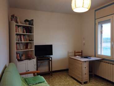 Ein gemütliches Zimmer hat 'ne grüne Couch, 'nen Holzschreibtisch, 'nen Stuhl, 'ne große Fensterfront, 'nen Bücherregal voller Bücher, 'nen Fernseher auf 'nem kleinen Ständer und 'ne warme Beleuchtung.