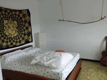 Ein gemütliches Schlafzimmer mit einem ungemachten Bett, einer gemusterten Tagesdecke, einem kunstvollen Wandteppich, einem hängenden Ast, einer Stehlampe und einer Gitarre, die an der schlichten weißen Wand lehnt.