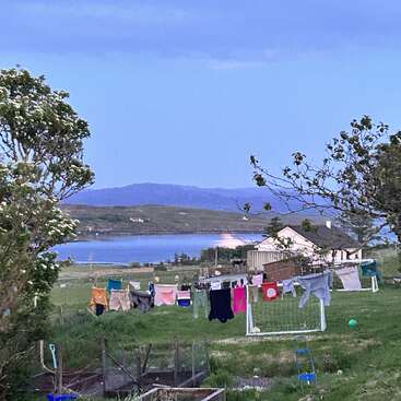 Das Bild zeigt eine ruhige ländliche Landschaft mit einem Haus, einer Wäscheleine und fernen Bergen vor einem blauen Himmel und einem ruhigen Gewässer.