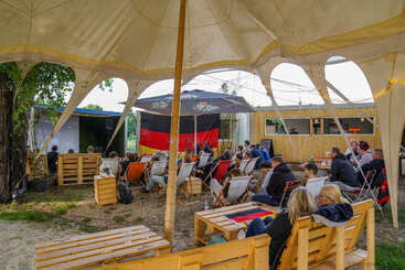 A imagem mostra um grupo de pessoas reunidas em um ambiente externo, cercado por uma cerca, com uma mesa e cadeiras visíveis ao fundo. A atmosfera parece ser descontraída e social.