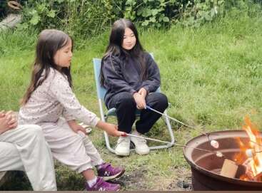 Dos niñas están sentadas junto a una hoguera en una zona cubierta de hierba, asando malvaviscos en palitos. Una está sentada en una silla y la otra, arrodillada a su lado.
