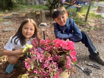 Dos niños están fuera, sonriendo junto a una gran cesta de flores de un rosa intenso. Cerca hay herramientas de jardinería. La luz del sol ilumina suavemente esta alegre escena natural.