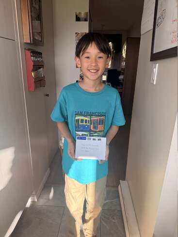 Un niño sonriente está de pie en el interior de una casa, sosteniendo un sobre. Lleva una camiseta azul de San Francisco y pantalones beige. La luz del sol ilumina su rostro. Al fondo se ve un pasillo.