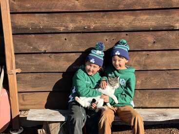 Dos niños sonrientes, vestidos con sudaderas verdes y gorros de invierno a juego, están sentados en un banco de madera, sosteniendo alegremente a un gato blanco y gris junto a una pared rústica de madera.