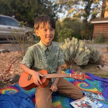 Un niño pequeño está sentado al aire libre sobre una manta de colores teñida al estilo tie-dye, sonriendo y con un ukelele en las manos. Lleva una camiseta verde y unos pantalones marrones. Cerca de él hay revistas tiradas por el suelo.