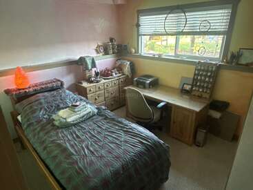 Una acogedora habitación con una cama individual, un escritorio de madera y una cómoda. Ventana con persianas, atrapasueños, una lámpara de sal, adornos y diversos objetos personales dispuestos con orden.