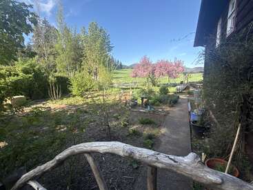 Una escena rústica de jardín con un sendero sinuoso, árboles en flor, plantas verdes, una barandilla de madera y una casa acogedora, todo ello bajo un cielo azul intenso.