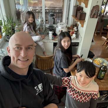 Un hombre alegre y tres niños están en una acogedora cocina, preparando la comida juntos. La cocina está llena de luz natural y se ven plantas y muebles de madera.