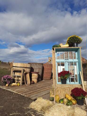 Une exposition d'automne rustique comprend des bottes de foin, des fleurs colorées, des courges, des tonneaux et un cadre de fenêtre réutilisé, le tout sous un ciel nuageux dramatique sur une plate-forme en bois.