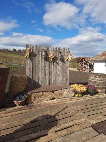Une scène de ferme rustique comprend une clôture en bois ornée de lanternes et de maïs, des bottes de foin, des tonneaux, des paniers de produits, des fleurs et un magnifique ciel bleu.