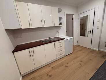 Esta imagem mostra uma cozinha pequena e moderna com armários em tom creme, bancada de madeira, pia, minigeladeira, armários superiores, prateleiras abertas e piso de madeira clara. Estilo minimalista.