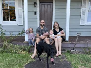 Uma família feliz de cinco pessoas, incluindo dois bebês, está sentada na varanda, com um cachorro preto na frente. A casa é cinza com acabamentos brancos.