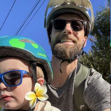 Um homem e uma criança, ambos usando capacete e óculos escuros, aproveitam um dia ensolarado ao ar livre. A criança tem uma flor amarela atrás da orelha.