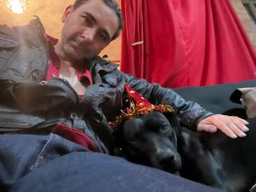 A man in a black jacket poses on a couch with a black dog wearing a festive party hat. Red curtains hang in the background, creating warmth.