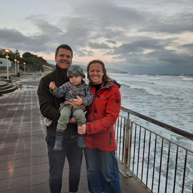 A happy family stands on a seaside promenade during sunset, smiling together. Waves crash against the shore, and dramatic clouds fill the sky above them.