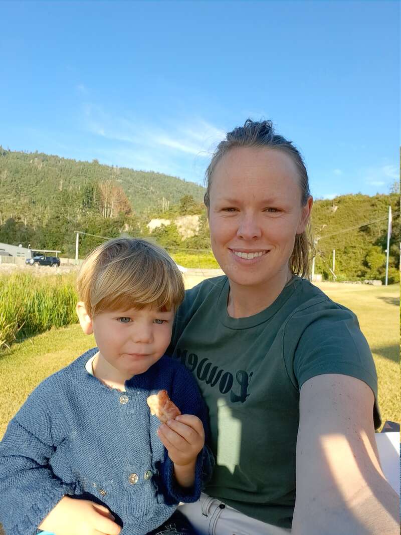 A woman and a young child are outdoors, sitting together on a sunny day. They both smile, with scenic green hills and blue sky in the background.