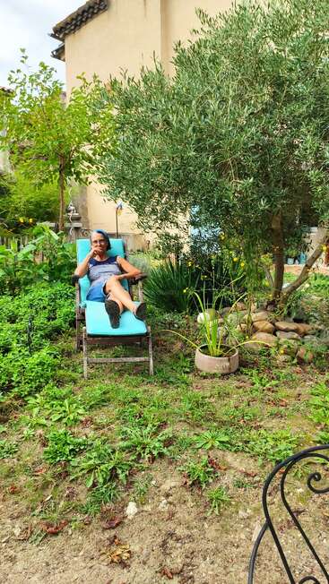 A woman relaxes on a turquoise lounge chair in a lush, green garden, surrounded by trees, plants, a potted aquatic plant, and a stone-bordered tree.