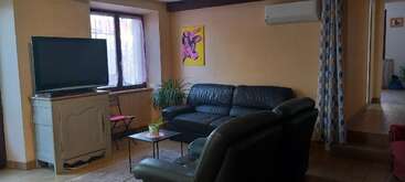 The cozy living room features black leather sofas, a TV, a small table with a plant, a window, a pop art painting, and beige walls.