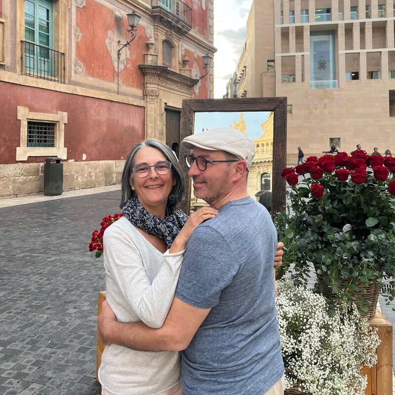 A smiling couple embraces in a charming European street, surrounded by red roses and white flowers, with historic buildings and a decorative mirror in the background.