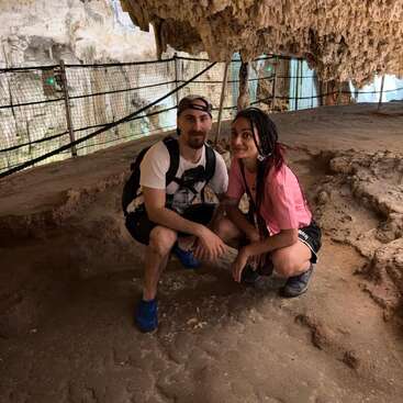 Um homem e uma mulher se agacham juntos, sorrindo dentro de uma caverna. As estalactites estão suspensas no alto, o solo rochoso abaixo e a cerca de segurança alinha o caminho da caverna ao fundo.