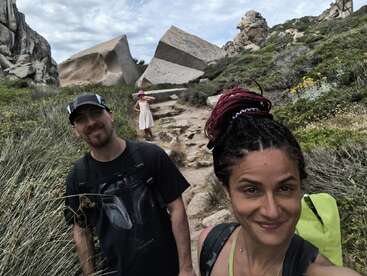 Um casal sorridente tira uma selfie durante uma caminhada. Atrás deles, uma criança de vestido está em um caminho rochoso perto de grandes formações de pedra de formato único.