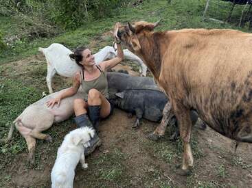 Eine Frau sitzt auf dem Boden, umgeben von Ziegen, Schweinen und einem Hund, und hebt die Hand zu einem High-Five, auf einer Wiese.