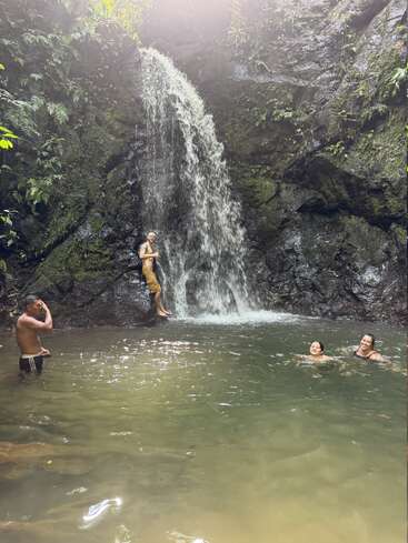 Vier Leute genießen ein erfrischendes Bad unter einem Wasserfall in einer üppig grünen, tropischen Umgebung, umgeben von Felsen und bunter Vegetation. Das Sonnenlicht scheint durch die Bäume.