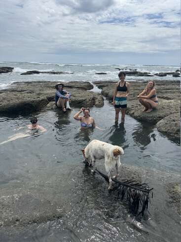 Fünf Leute und ein Hund chillen an einem natürlichen Gezeitentümpel am Meer. Einige sitzen oder schwimmen, andere stehen da, und das alles unter einem bewölkten Himmel und umgeben von Felsen.