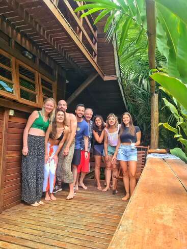 Un grupo de ocho personas sonrientes posan descalzas en una terraza de madera rodeada de exuberantes plantas tropicales y una rústica cabaña de madera, irradiando alegría y relajación.
