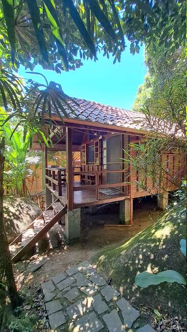 Una encantadora cabaña de madera con tejado de tejas está enclavada entre exuberantes plantas tropicales, con escalones de piedra que conducen a su porche abierto.