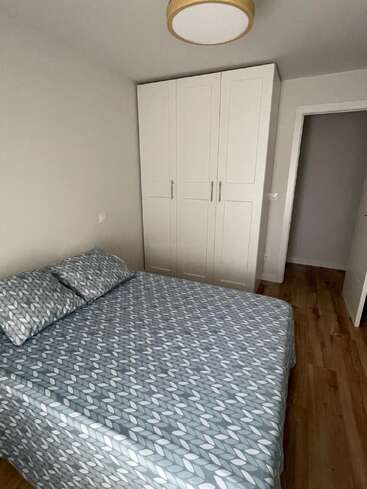 Una habitación pequeña y sencilla con una cama doble con una colcha de motivos geométricos, dos almohadas, un gran armario blanco, suelo de madera y paredes de color neutro.