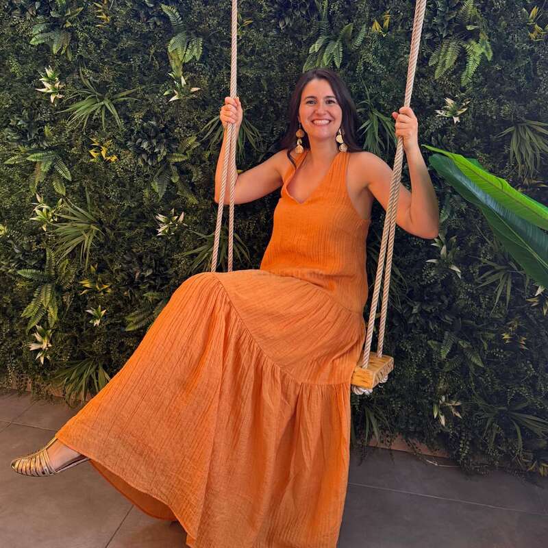 Una mujer con un vestido naranja está sentada sonriendo en un columpio, con una pared cubierta de hojas verdes y un letrero de neón detrás de ella. El letrero anuncia Schweppes en español.
