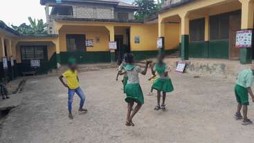 Varios niños están jugando en el patio de un colegio. El edificio es de color amarillo y verde, y tiene carteles en las paredes. El ambiente es animado y alegre.