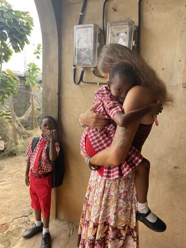 Una mujer abraza a un niño sonriente que lleva un uniforme escolar a cuadros rojos. Otro niño, vestido de forma similar, está de pie cerca, observando. La escena irradia calidez y cariño.