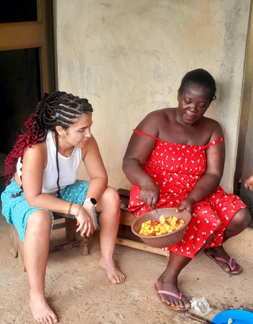 Dos mujeres están sentadas juntas frente a una casa. Una, vestida de rojo, prepara la comida en un cuenco, mientras que la otra, vestida de azul, la observa con atención, compartiendo ese momento.