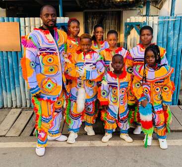 Un grupo de personas, entre las que hay niños y adultos, visten vibrantes y coloridos trajes tradicionales con intrincados dibujos, permanecen juntas y sonrientes en una calle de la ciudad.