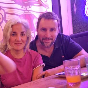 A smiling man and woman sit closely together at a table in a restaurant with colorful lighting, drinks, and vibrant wall art in the background, enjoying their time.