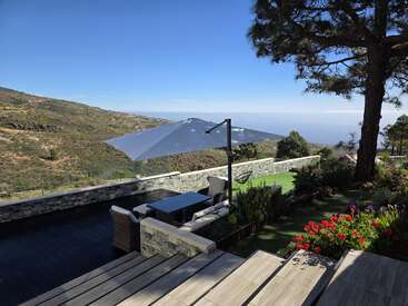 A beautiful terrace with seating, a large umbrella, and vibrant flowers overlooks a scenic mountain landscape under a clear blue sky, offering peaceful relaxation.