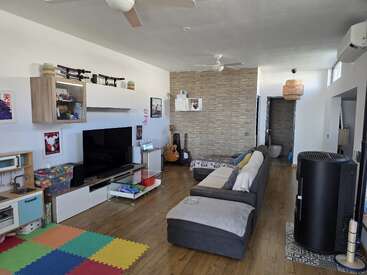 Modern living room with wooden flooring, sectional sofa, TV, guitars, play area with colorful mats, ceiling fans, wall art, and natural light. Cozy and organized space.