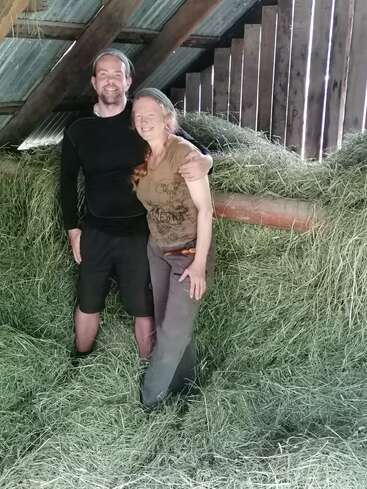 A smiling man and woman stand close together in a barn loft, surrounded by piles of hay, under a slanted metal and wooden roof.