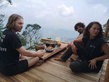Three people sit barefoot on a wooden deck, sharing food on a small table, surrounded by lush trees and mountains under a partly cloudy sky. Relaxing, serene atmosphere.