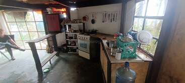 This rustic kitchen features open shelves, a stove, hanging utensils, a dish rack by the window, a large water jug, and a person sitting nearby.