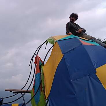 A man sits atop a colorful, geometric dome structure against a cloudy sky. The dome is patched with yellow, blue, green, and orange sections, looking playful.