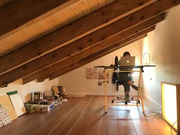 Das Bild zeigt eine Person, die an einem Schreibtisch in einem Raum mit Holzboden und Deckenbalken sitzt, umgeben von verschiedenen Gegenständen auf dem Boden und an den Wänden.