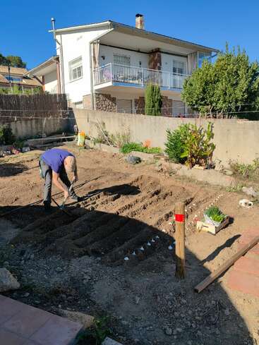 Ein Mann gräbt mit einer Schaufel in einem Garten, umgeben von Pflanzen und einem Haus im Hintergrund an einem sonnigen Tag.