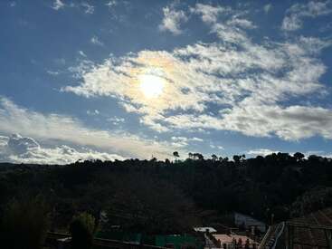 Das Bild zeigt eine ruhige Landschaft mit einem blauen Himmel mit weißen Wolken, einer strahlenden Sonne und einem Berghang mit Bäumen und Gebäuden in der Ferne.