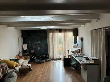 Das Bild zeigt einen Raum mit weißen Wänden und einer weißen Decke, in dem ein Schreibtisch, ein Stuhl und ein Fenster neben einer Couch und einer Tafel stehen, mit einem Holzboden und verschiedenen verstreuten Gegenständen.