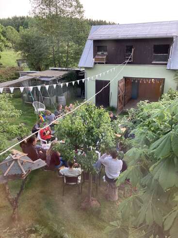 Un grupo de personas se reúne al aire libre en un frondoso jardín, sentadas alrededor de unas mesas cerca de un edificio verde, disfrutando de una fiesta de verano con adornos y árboles.